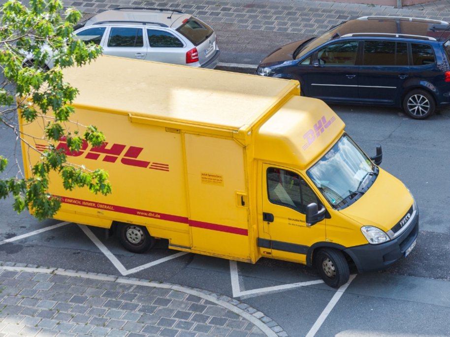 Ein gelber DHL-Lieferwagen parkt auf einem asphaltierten Parkplatz neben zwei Personenwagen, mit einem Baum im Vordergrund.