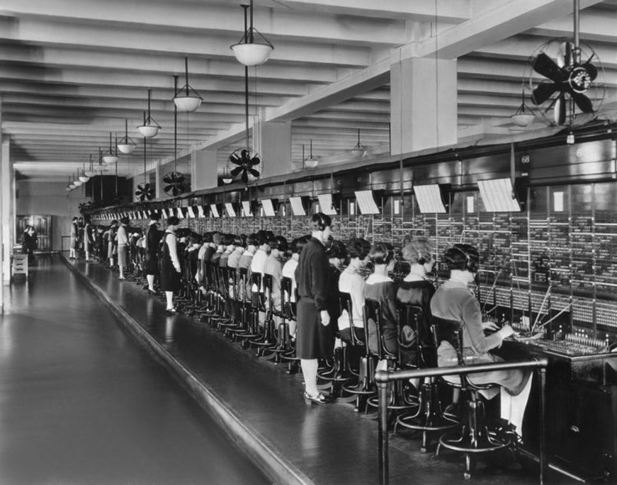 Ein historisches Schwarz-Weiß-Foto zeigt eine große Telefonzentrale mit vielen Telefonistinnen, die an einer langen Schaltwand sitzen und Anrufe verbinden. Eine Aufseherin steht daneben und überwacht die Arbeit.