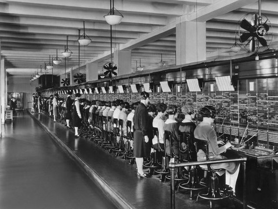Ein historisches Schwarz-Weiß-Foto zeigt eine große Telefonzentrale mit vielen Telefonistinnen, die an einer langen Schaltwand sitzen und Anrufe verbinden. Eine Aufseherin steht daneben und überwacht die Arbeit.