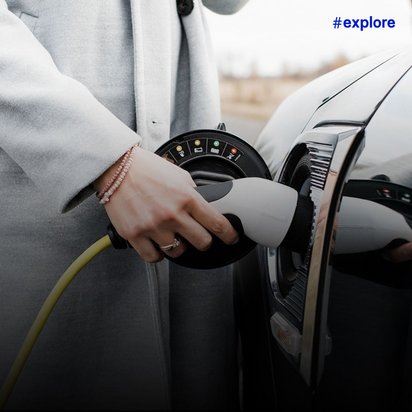 Eine Hand in hellgrauem Mantel mit Armband steckt einen Ladestecker in die Ladebuchse eines Elektroautos, erkennbar am gelben Ladekabel und dem speziellen Anschluss.