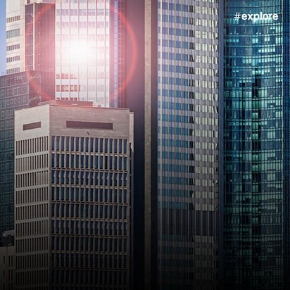 Eine Stadtansicht mit mehreren Wolkenkratzern und Bürogebäuden in unterschiedlichen Höhen und Materialien. Die Sonne scheint zwischen den Gebäuden hindurch und erzeugt einen hellen Lichteffekt.