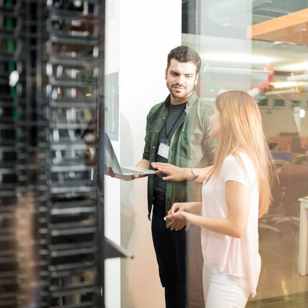Zwei IT-Fachkräfte stehen im Serverraum und besprechen Daten auf einem Laptop vor einem Serverschrank