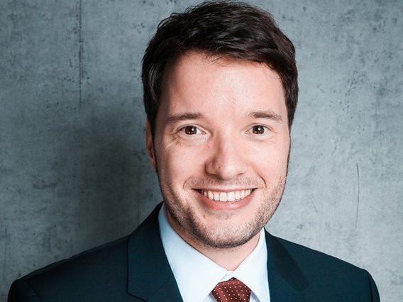 Ein professionelles Porträt von Manuel Hagemann. Er trägt einen dunklen Anzug, ein weißes Hemd und eine rote Krawatte, mit einem strukturierten grauen Hintergrund.