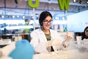 Eine junge Person in einem weißen Laborkittel führt ein chemisches Experiment durch. Sie trägt eine Brille und arbeitet konzentriert mit Laborausrüstung.