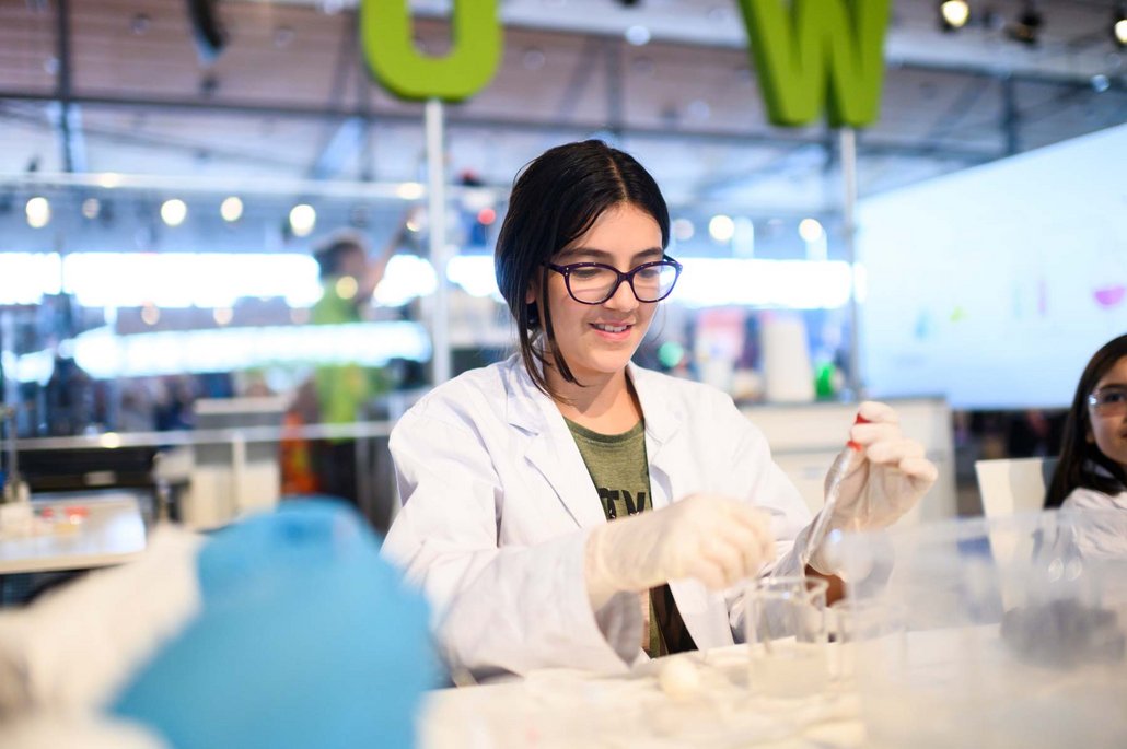 Eine junge Person in einem weißen Laborkittel führt ein chemisches Experiment durch. Sie trägt eine Brille und arbeitet konzentriert mit Laborausrüstung.