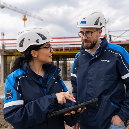Zwei TÜV NORD-Mitarbeitende mit Helmen und Tablets im Gespräch auf einer Baustelle beim baubegleitenden Qualitätscontrolling