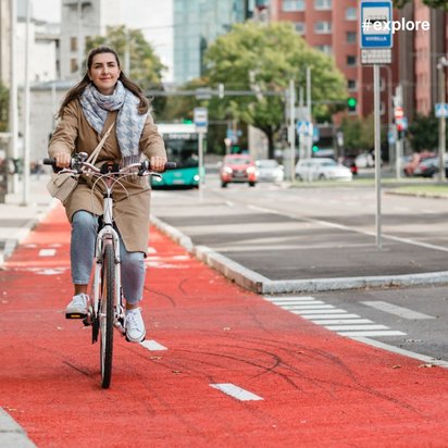 Lächelnde Frau in beigem Mantel und Schal fährt Fahrrad auf rotem Radweg in städtischer Umgebung.