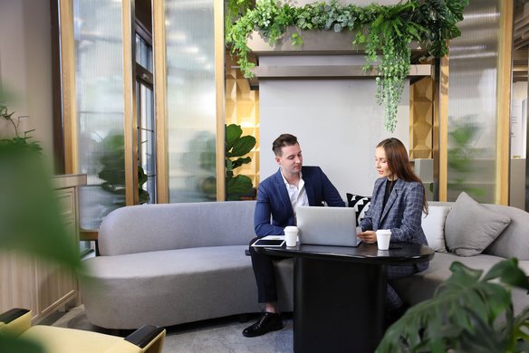 Zwei Geschäftsleute sitzen mit Laptop und Kaffee in einer modernen Lounge mit Pflanzen an einem Tisch.