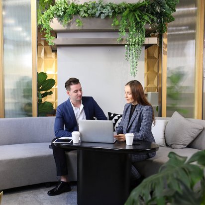 Zwei Geschäftsleute sitzen mit Laptop und Kaffee in einer modernen Lounge mit Pflanzen an einem Tisch.
