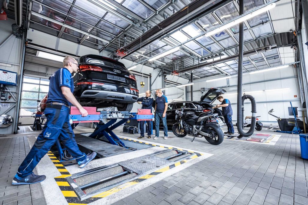  TÜV NORD Mitarbeiter prüfen Autos und Motorräder in einer modernen, hellen Werkstatthalle mit Hebebühne.