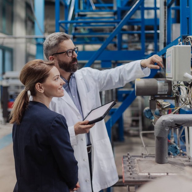 Ein technischer Experte erklärt einer Kollegin in einer Industrieanlage die Mess- und Kontrollsysteme. Beide stehen vor einem Schaltschrank, im Hintergrund sind Rohre und metallische Strukturen sichtbar.