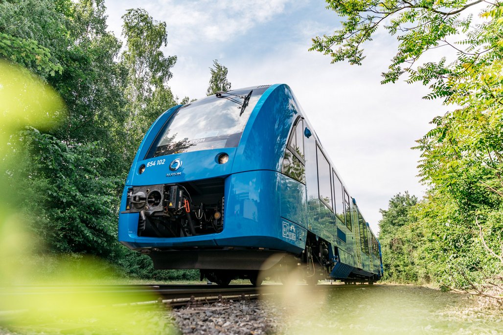 Ein blauer Personenzug mit der Nummer 854 102 fährt durch eine grüne Waldlandschaft auf Schienen. Die moderne Zugfront hat eine aerodynamische Form und große Fenster, umgeben von üppiger Vegetation.