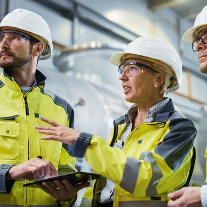 Drei Fachkräfte in Warnkleidung und Schutzhelmen prüfen gemeinsam technische Daten auf einem Tablet in einer Industrieumgebung.