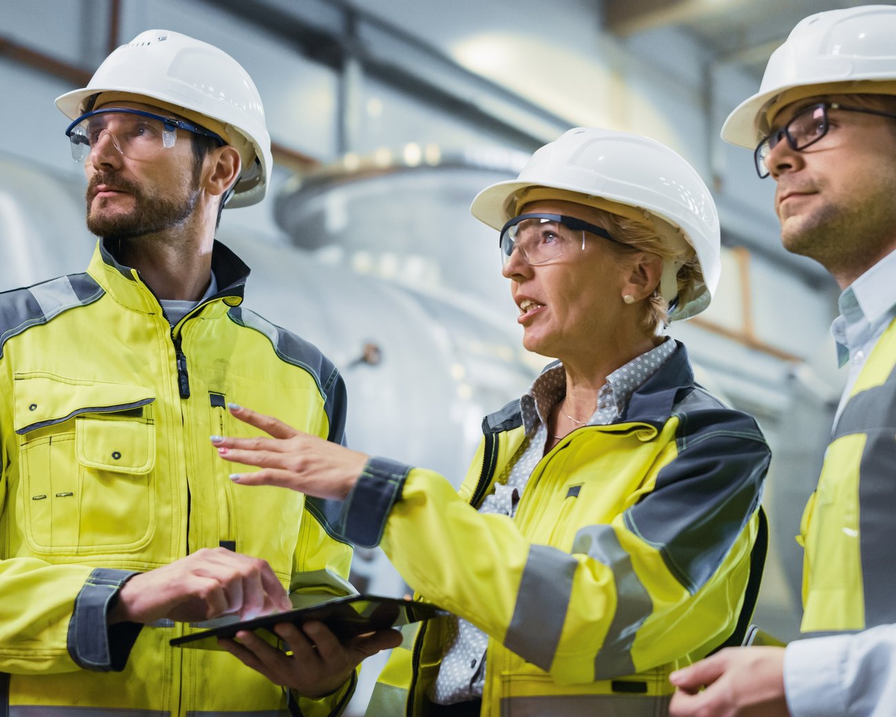 Drei Fachkräfte in Warnkleidung und Schutzhelmen prüfen gemeinsam technische Daten auf einem Tablet in einer Industrieumgebung.