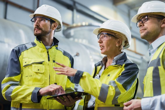 Drei Fachkräfte in Warnkleidung und Schutzhelmen prüfen gemeinsam technische Daten auf einem Tablet in einer Industrieumgebung.