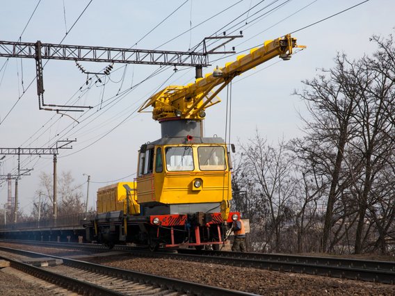 Gelbes Bahndienstfahrzeug mit Kranaufsatz auf Eisenbahngleisen unter Oberleitungen, Bäume und Strommasten im Hintergrund.