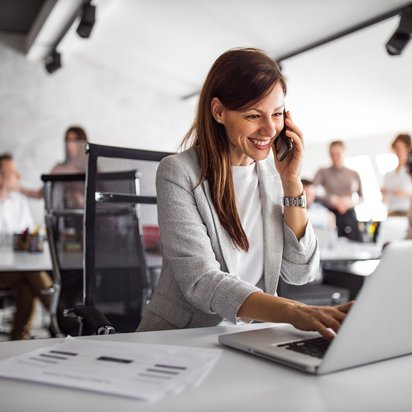 Transfergesellschaft von TÜV NORD Transfer – Unterstützung bei beruflicher Neuorientierung Mitarbeiterin am Telefon, die an einem Laptop arbeitet und in einem modernen Büroumfeld sitzt