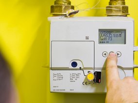 Finger drückt Taste an weißem digitalen Gaszähler mit LCD-Display, das Verbrauchsdaten und Tarif anzeigt.