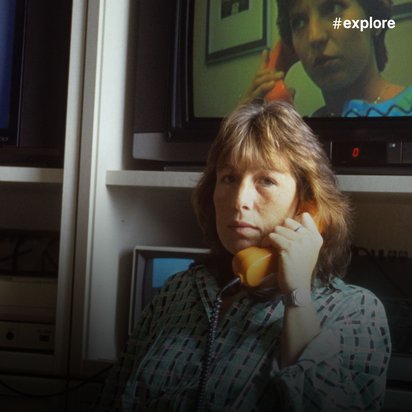 Frau telefoniert mit gelbem Hörer vor mehreren Röhrenfernsehern, die Bildschirmtext ("Bildschirmtext") anzeigen. Die Szene stammt vermutlich aus den 1980er Jahren.