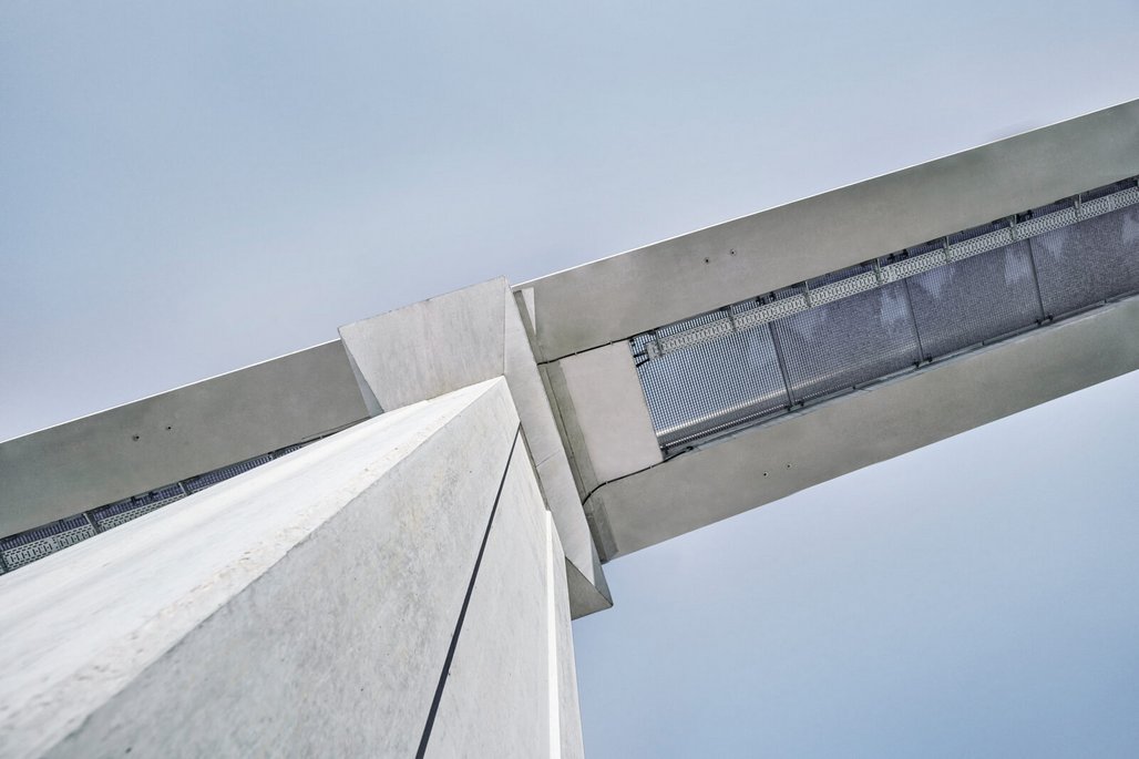 Aufnahme von unten zeigt eine moderne Betonbrücke mit Metallgeländer, die sich diagonal über einen bewölkten Himmel erstreckt. Die minimalistische Architektur betont klare Linien und geometrische Formen in Grau- und Weißtönen.