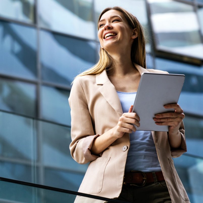 Frau in beigem Blazer mit Tablet vor einem modernen Glasgebäude.