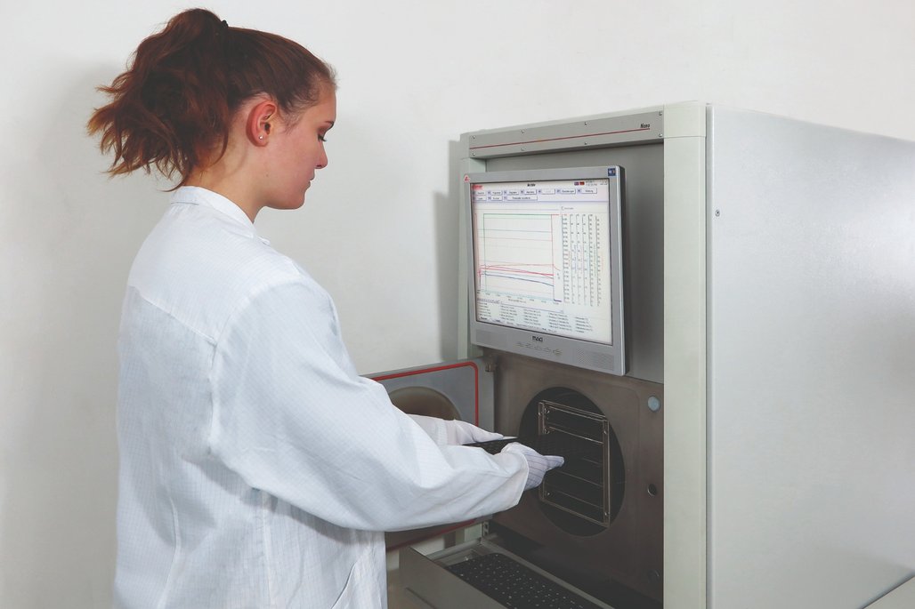 Eine Person in einem Laborkittel arbeitet mit einem Analysegerät. Der Bildschirm zeigt grafische Daten und Diagramme. Die Person trägt Handschuhe und führt eine Probe in das Gerät ein.