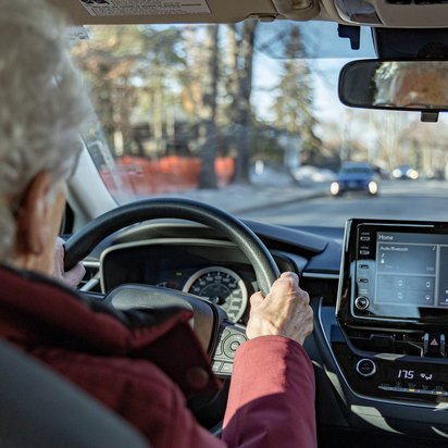 Ältere Person mit weißem Haar fährt ein Auto auf einer Stadtstraße bei Tageslicht