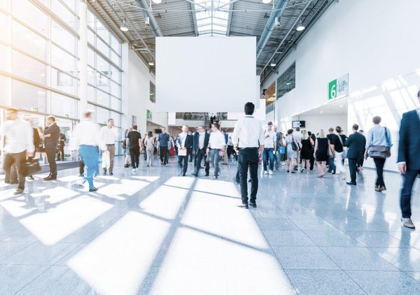 Bild von Menschen auf einer Messe oder Veranstaltungshalle mit vielen Ständen und heller Beleuchtung.