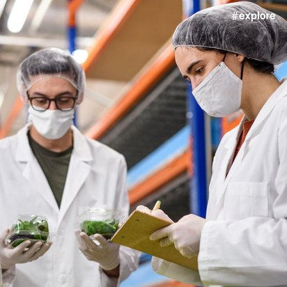 Zwei Personen in weißen Kitteln und Hauben halten grüne Pflanzen in Behältern und notieren etwas auf einem Klemmbrett in einem Lagerraum