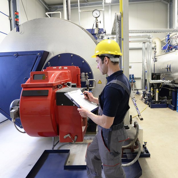 Techniker mit Schutzhelm prüft Industrieanlage mit roter Maschine und macht Notizen auf einem Klemmbrett