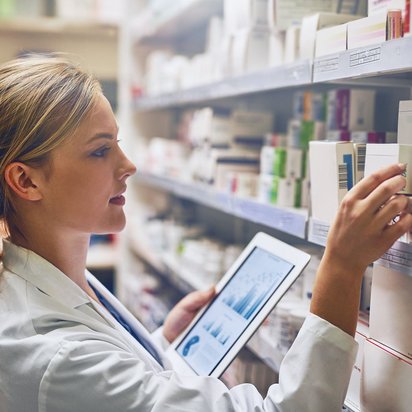 Apothekerin mit Tablet entnimmt Medikamentenschachtel aus einem Regal in der Apotheke.