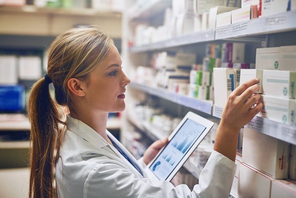 Apothekerin mit Tablet entnimmt Medikamentenschachtel aus einem Regal in der Apotheke.