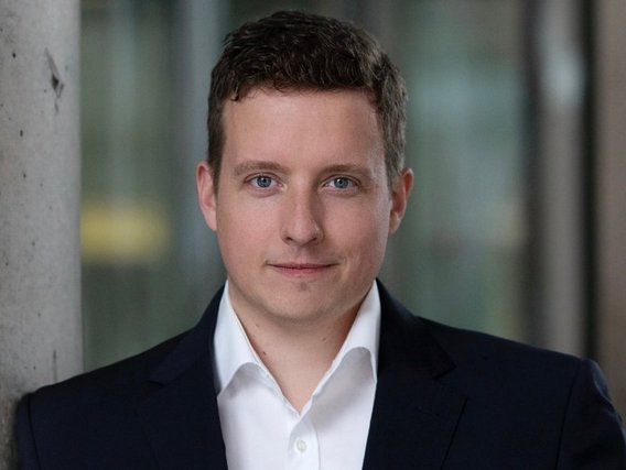 Malte Hübner in dunklem Anzug und weißem Hemd steht vor einem Bürogebäude. Er posiert für ein professionelles Porträtfoto neben einer Betonsäule mit unscharfem Hintergrund.