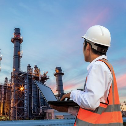 An officer in safety clothing stands next to an industrial plant with a laptop in his hands.