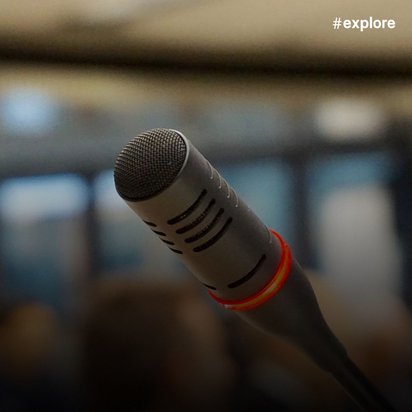 Nahaufnahme eines Mikrofons mit charakteristischem Gitterkopf, bereit für einen Sprecher mit verschwommenem Publikum im Hintergrund.