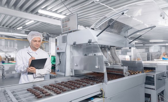 Mitarbeiter:in in Schutzkleidung prüft mit Tablet die Schokoladenproduktion an einer Fertigungsstraße.