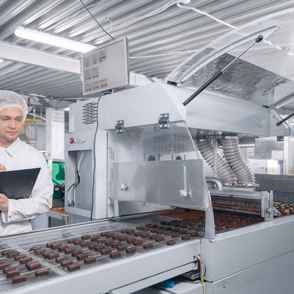 Mitarbeiter:in in Schutzkleidung prüft mit Tablet die Schokoladenproduktion an einer Fertigungsstraße.