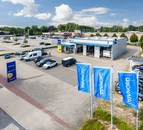 TÜV NORD Prüfstation mit großem Parkplatz, mehreren Autos und drei blauen TÜV NORD Flaggen im Vordergrund.