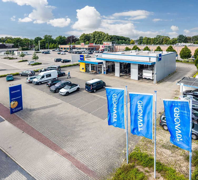 TÜV NORD Prüfstation mit großem Parkplatz, mehreren Autos und drei blauen TÜV NORD Flaggen im Vordergrund.