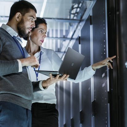 Zwei Personen arbeiten mit einem Laptop zwischen Serverracks in einem Rechenzentrum. Das Bild symbolisiert Eignungsdiagnostik und Auswahlverfahren für sicherheitsrelevante Funktionen.