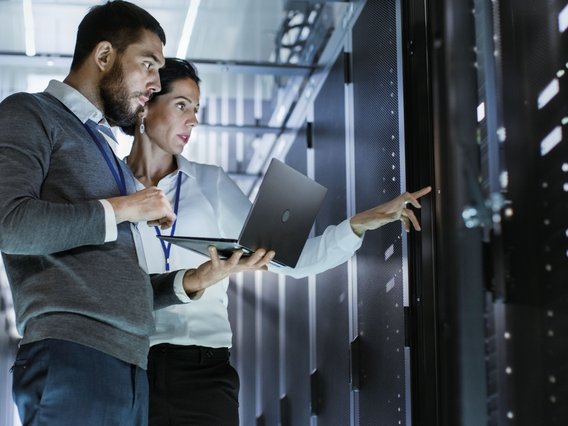 Zwei Personen arbeiten mit einem Laptop zwischen Serverracks in einem Rechenzentrum. Das Bild symbolisiert Eignungsdiagnostik und Auswahlverfahren für sicherheitsrelevante Funktionen.