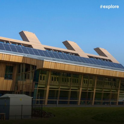 Nachhaltiges Gebäude mit Holzfassade, großer Glasfront und Solaranlage auf dem Dach, vier charakteristische Schornsteine und eine Grünfläche im Vordergrund.