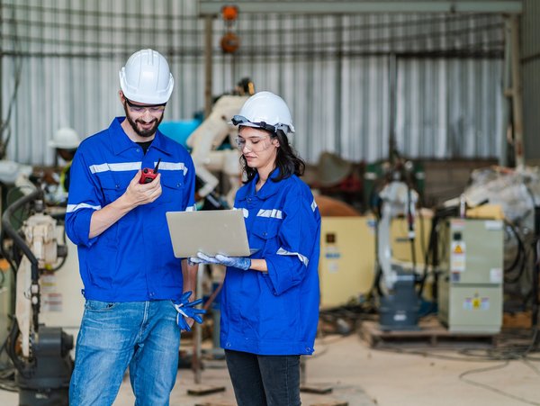 Zwei Fachleute arbeiten in einem Werk und führen eine Gefährdungsbeurteilung mit Laptop und Kommunikationsgerät durch