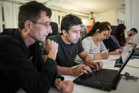 Mehrere Personen konzentrieren sich auf ihre Laptops in einer Schulungsumgebung, eine Person erklärt etwas.