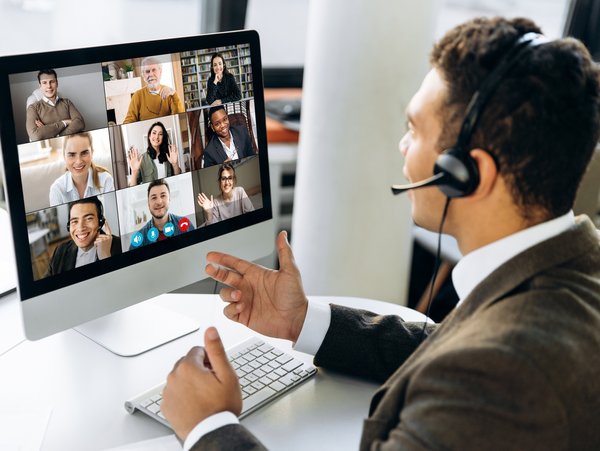 Mann hält eine Videokonferenz mit mehreren Teilnehmern die auf dem Computermonitor zu sehen sind