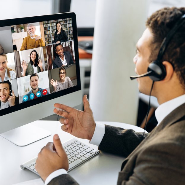 Mann hält eine Videokonferenz mit mehreren Teilnehmern die auf dem Computermonitor zu sehen sind