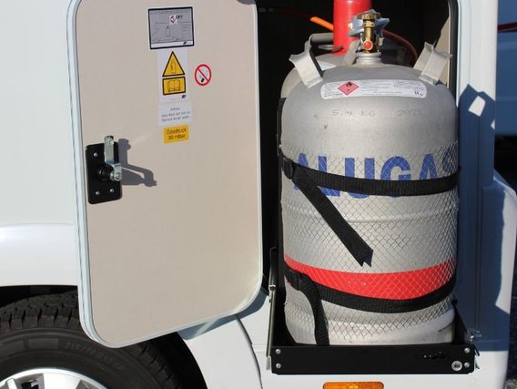 Graue Gasflasche mit blauen "ALUGAS"-Markierungen, sicher in einer Fahrzeughalterung montiert. Warnhinweise auf der Türinnenseite.