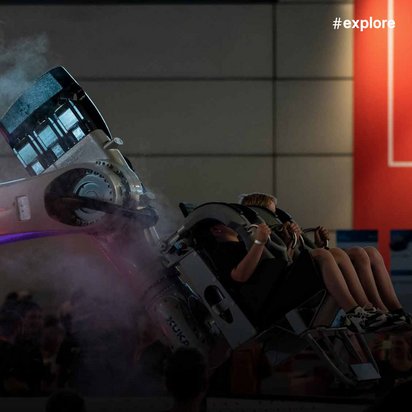 Eine beeindruckende Roboterinstallation, möglicherweise ein KUKA-Industrieroboter, der eine Art Fahrgeschäft oder Simulator betreibt. Einige Personen sitzen in einer von dem Roboterarm bewegten Gondel. Im Hintergrund ist das "Ideen Expo 19" Logo auf einer roten Wand zu sehen.