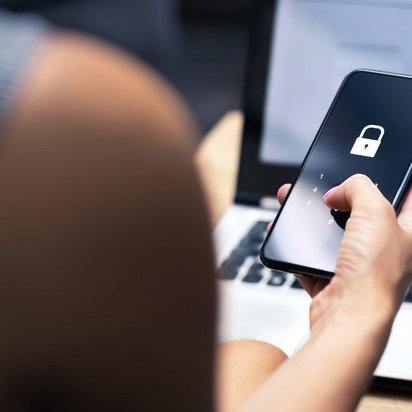 Eine Person hält ein Smartphone mit einem Schloss-Symbol auf dem Bildschirm, während im Hintergrund ein Laptop zu sehen ist.