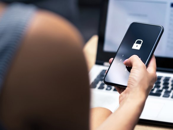Eine Person hält ein Smartphone mit einem Schloss-Symbol auf dem Bildschirm, während im Hintergrund ein Laptop zu sehen ist.